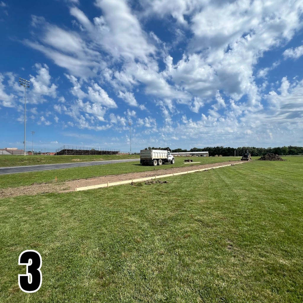 Green field with a road, a truck, and a pile of dirt. The sky is blue with clouds.