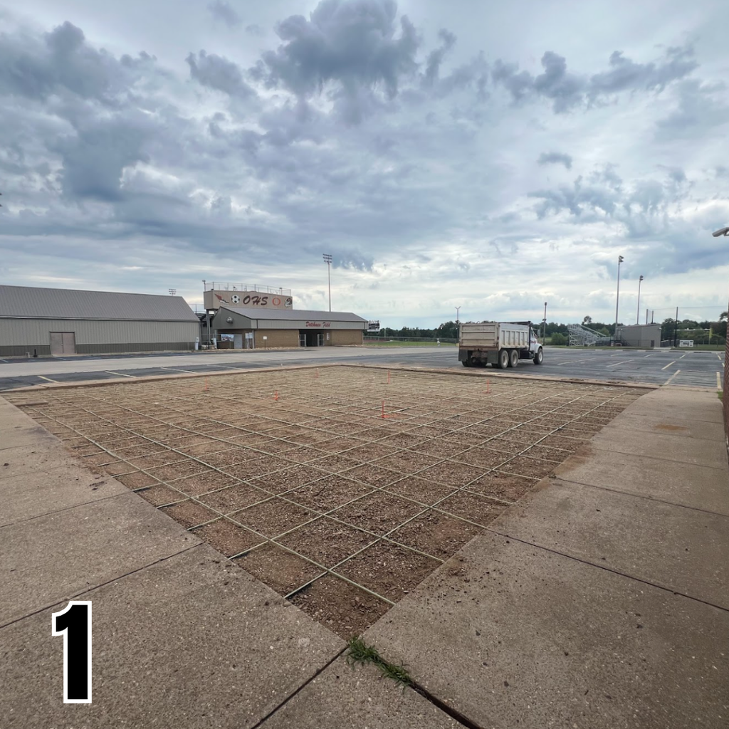 A large open area with a grid pattern in the middle, bordered by concrete walkways. A truck is visible on a road in the background.