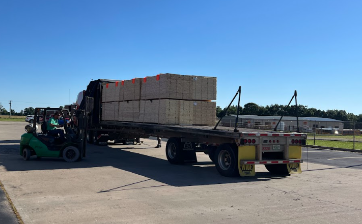 A forklift unloads wooden boxes from a trailer on a paved road, against a blue sky and distant trees.