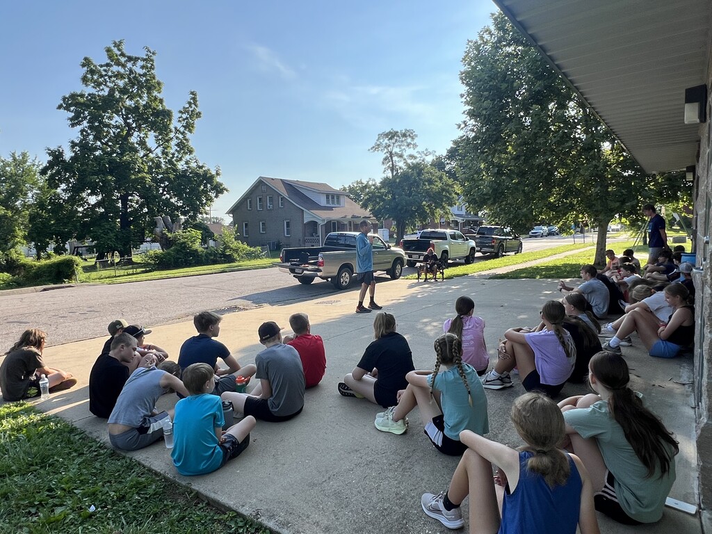 A group of students sit in a semicircle on a concrete sidewalk while a teacher stands in front of them.