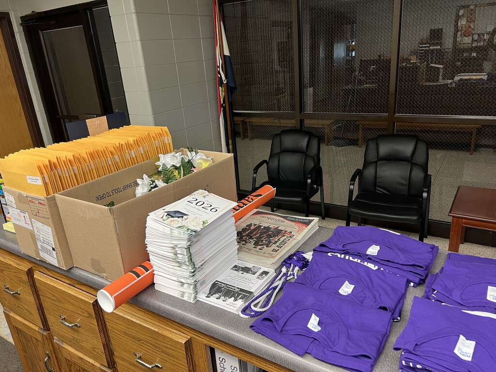 A counter with stacks of purple t-shirts, a box, books, and other items. Two black chairs and a glass wall are behind the counter.