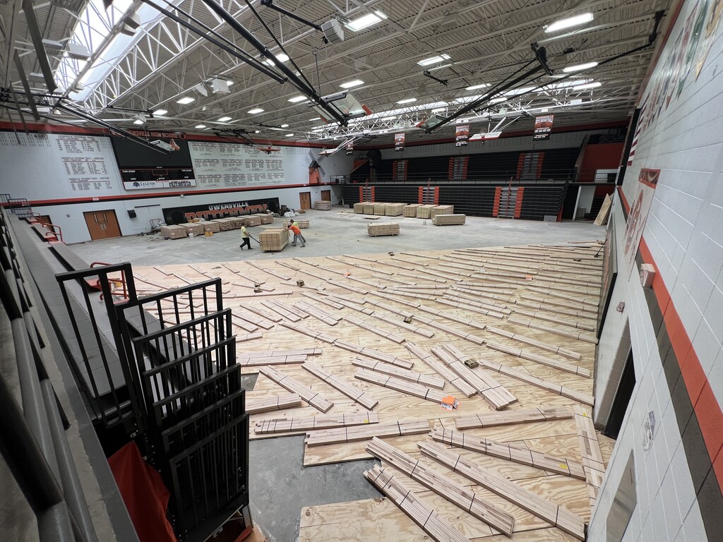 Interior of a large, open space with wooden flooring being installed. Bright lighting and metal supports are visible.