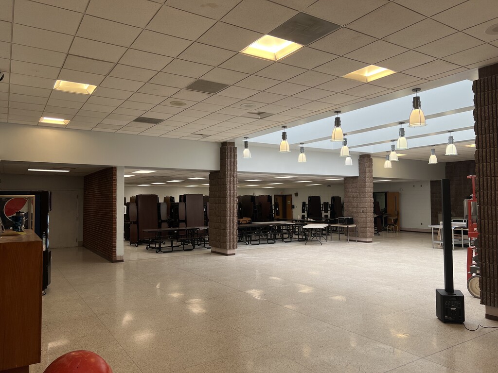 A large, empty room with white walls and tiled floor. The ceiling has square tiles and hanging lights.