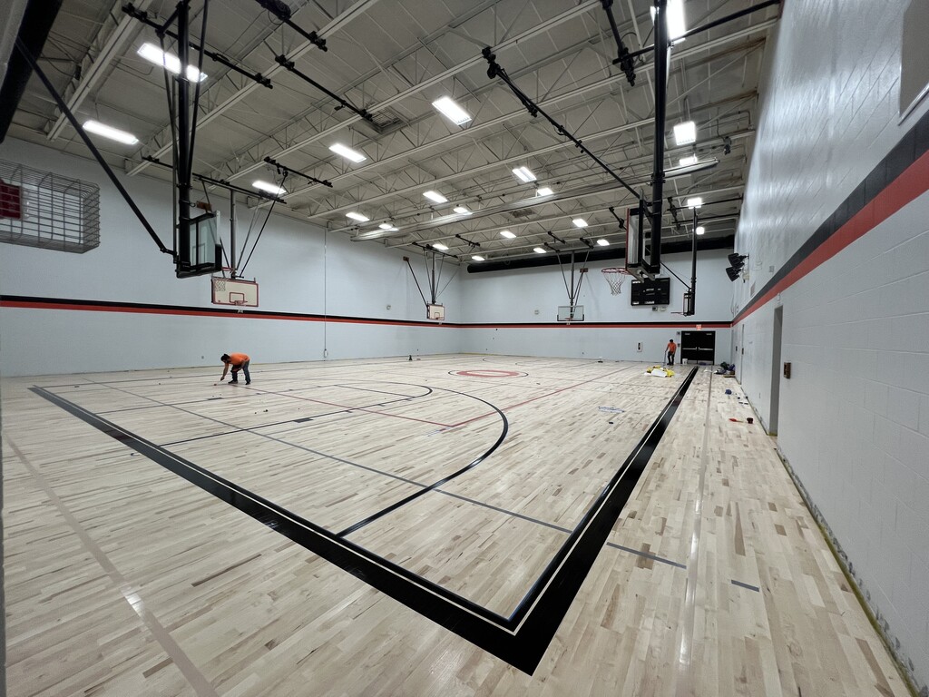 Indoor basketball court with a person working on the floor. White walls, red trim, and wooden flooring.