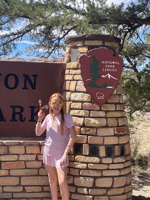 A girl in a pink outfit holds Flat Dutchy in front of a stone sign for Zion National Park.