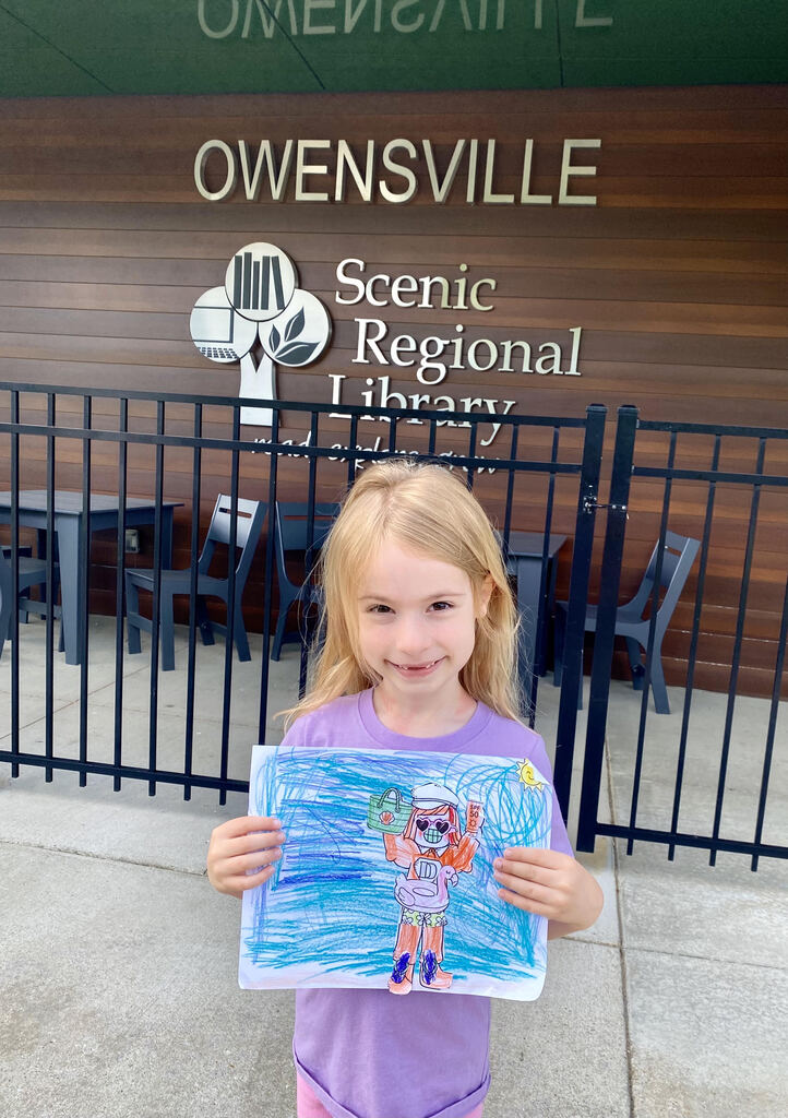 Child holds a drawing outside the Scenic Regional Library, smiling. Behind them, a black fence and tables are visible.