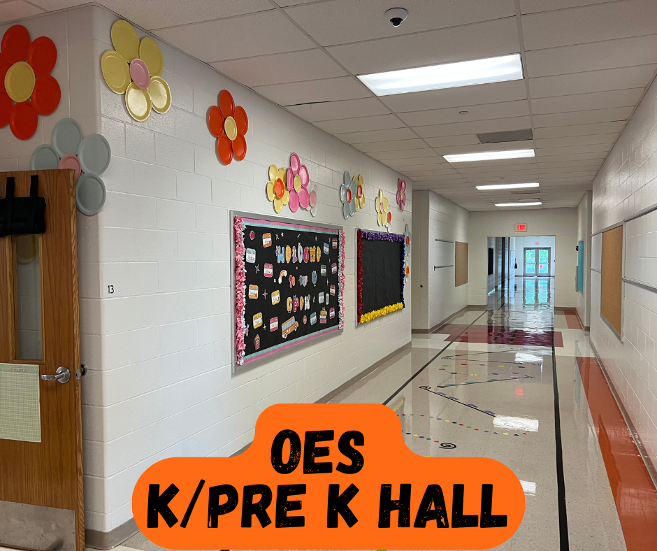A school hallway with white walls and orange trim. Flower decorations and bulletin boards adorn the walls. Bright lighting is overhead.