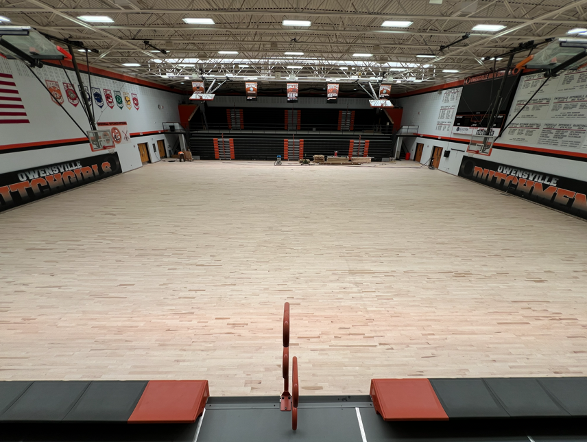 Large indoor basketball court with new hardwood flooring, orange and black walls, and a flag at the far end.