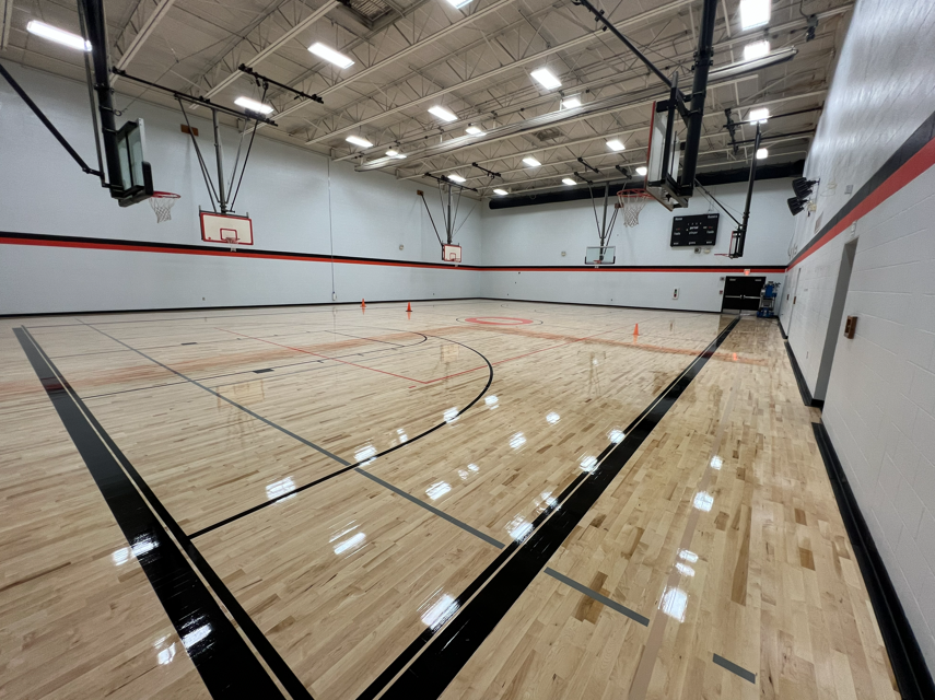 An empty indoor basketball court with polished wooden floors, black lines, and orange cones. The walls have red and white stripes, and there are basketball hoops and lights.
