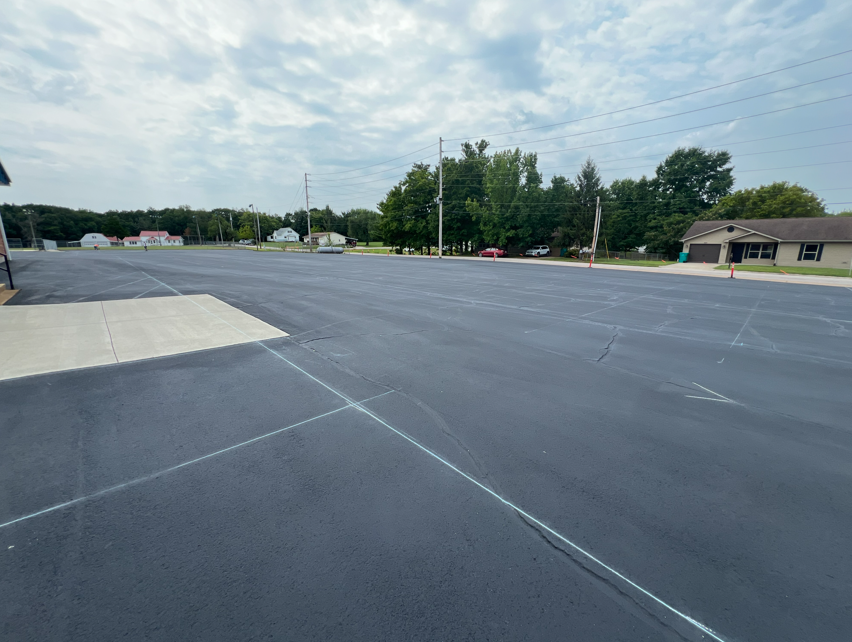 Wide, empty parking lot with freshly paved asphalt, white markings, and a small curb. Trees and houses visible in the background.