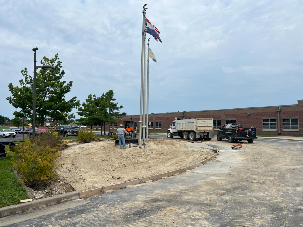 A person stands in a sandy area while construction vehicles are parked. A building is in the background.