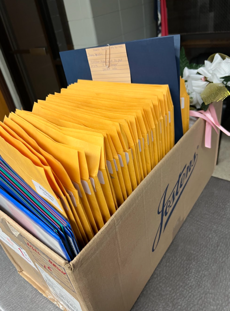 A box filled with yellow envelopes and other documents, including a blue folder, on a gray surface.