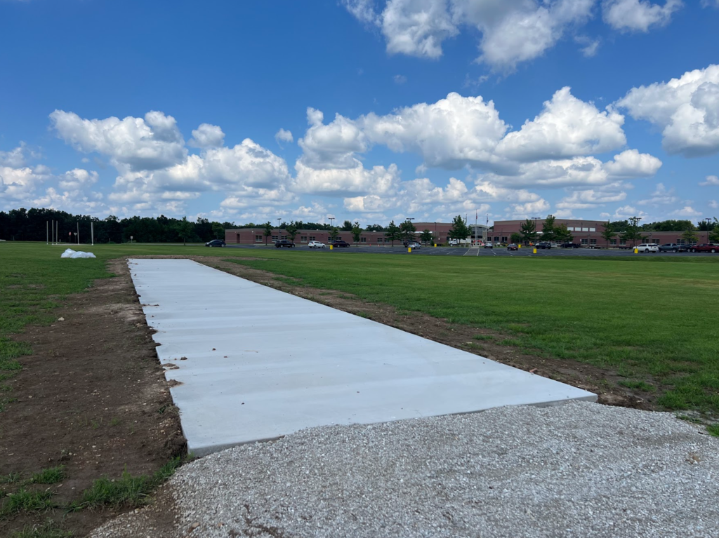 A concrete path extends across a grassy field with a blue sky and scattered clouds. Buildings in the background.