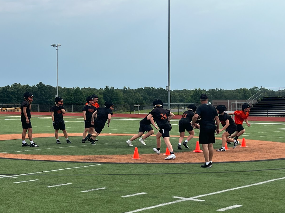 A group of individuals in black uniforms running on a football field. Some wear orange cones. Trees are in the background.
