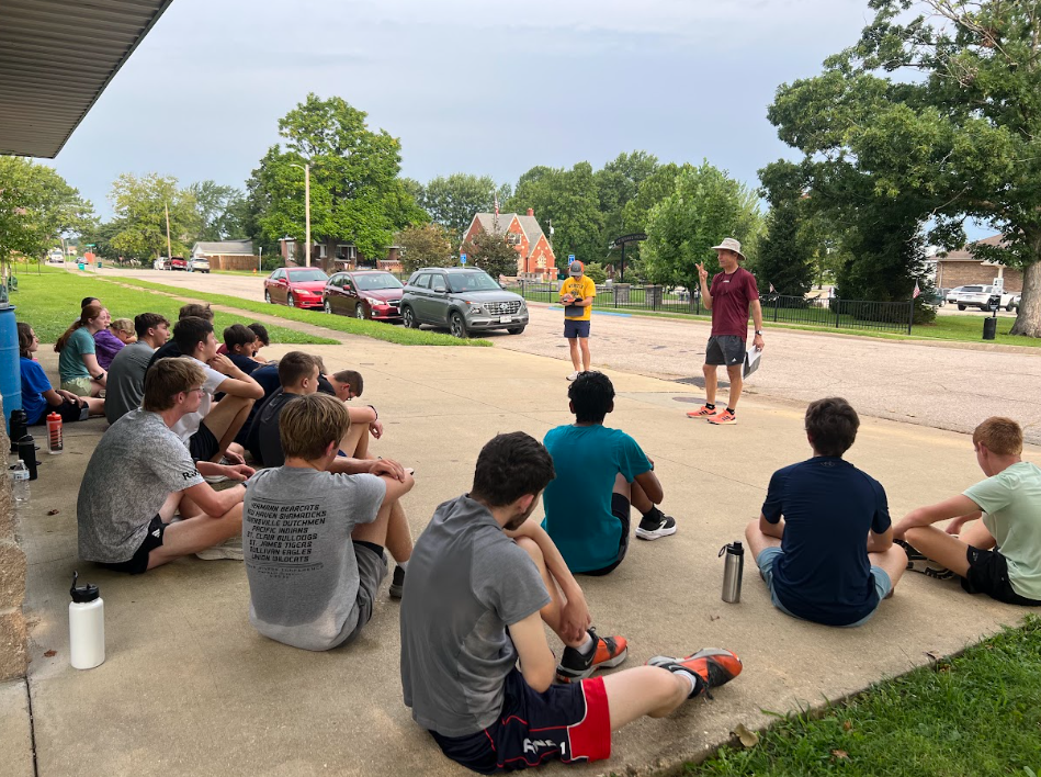 A group of people in casual wear sit on a sidewalk. One man stands in front of them. Trees and parked cars line the street.