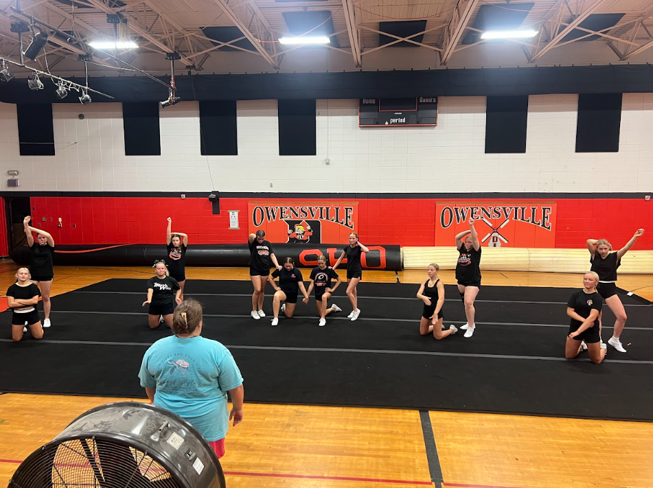 Group of people in black attire practicing cheerleading in a gym with red and white walls.