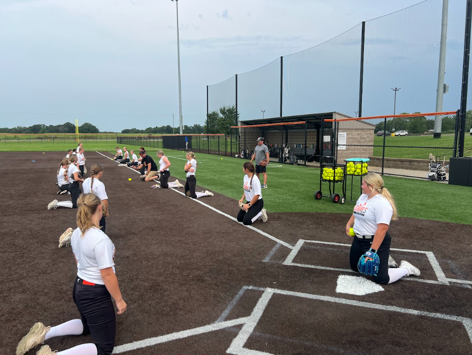 A group of people in white shirts and black pants practicing softball on a field with a net.