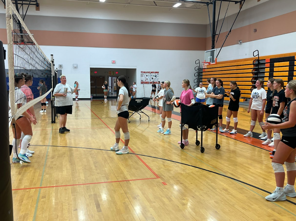An indoor volleyball court with several people. Some individuals are standing in groups while others are near the net.