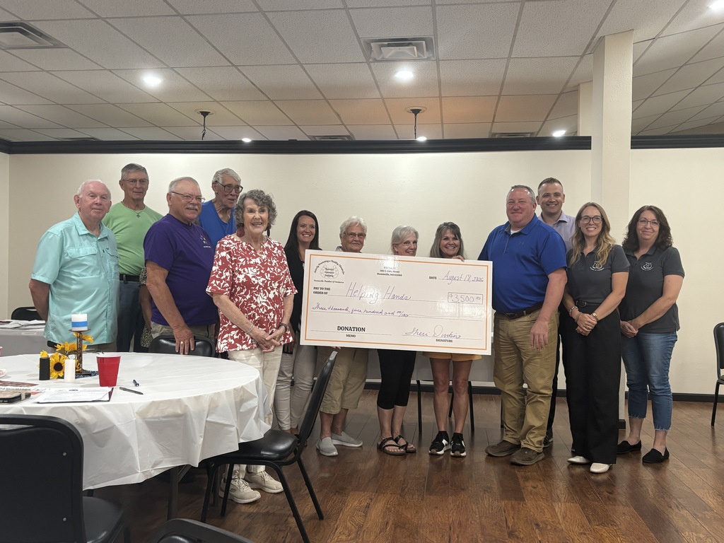 A group of people stand behind a table, presenting a large check to a woman in a floral top.