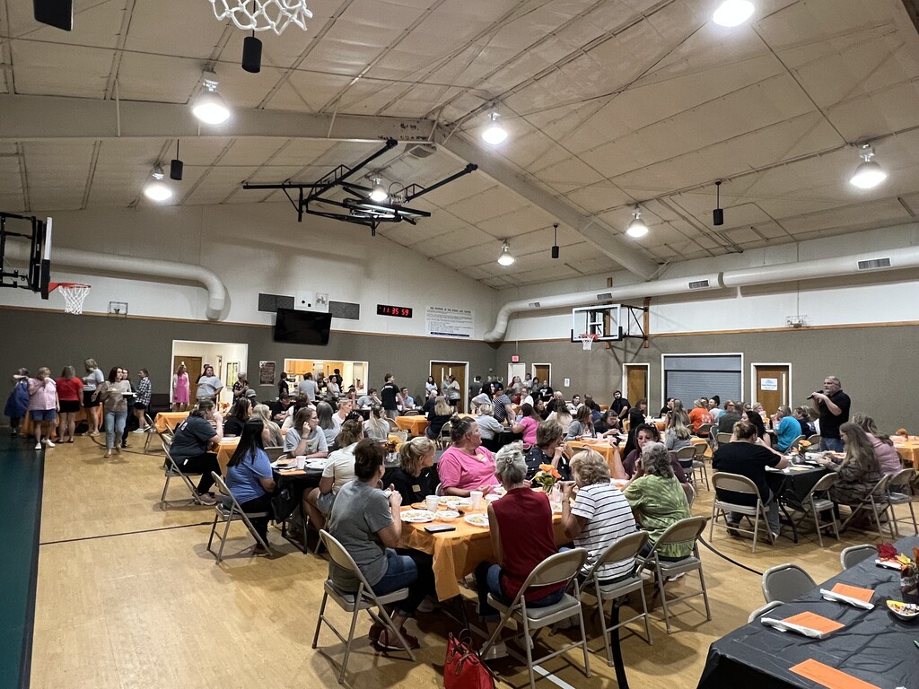 Many people sit at tables in a large room with wooden floors, basketball hoops, and lights.