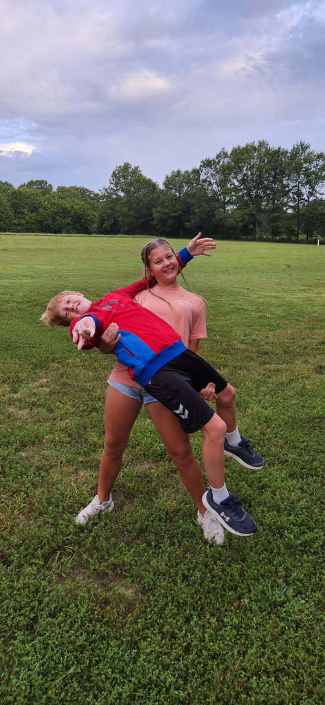Two individuals stand on a grassy field. One holds a child on their back. The child wears a red shirt. Trees surround the area.