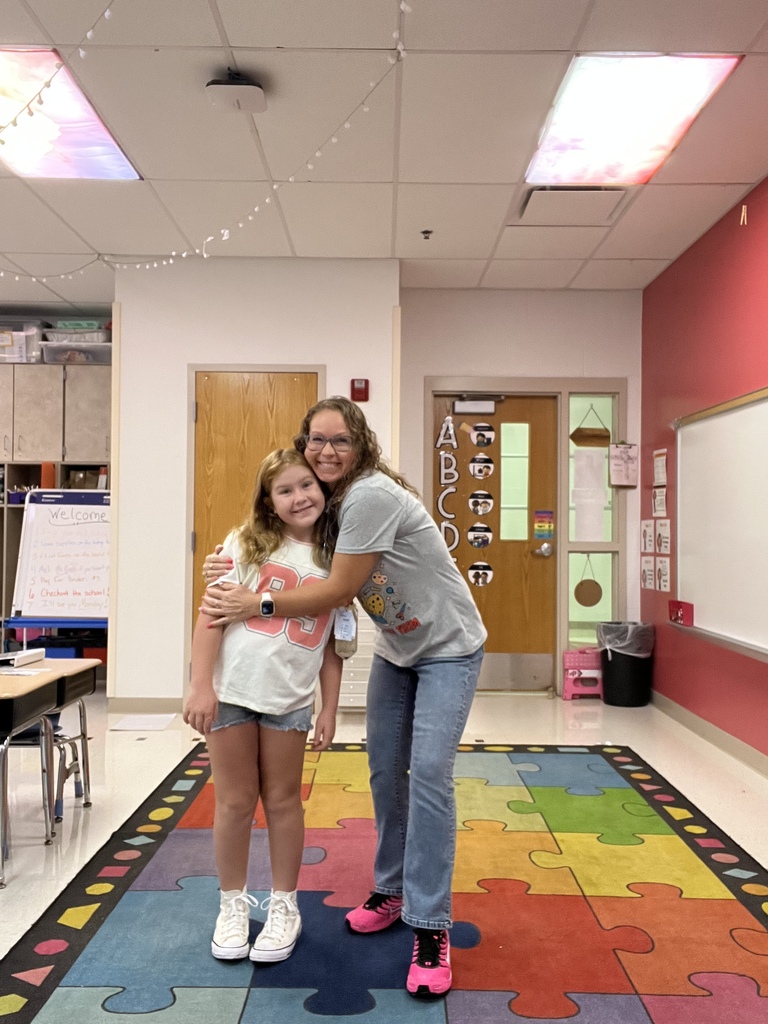A teacher and student stand in a classroom, embracing, near a colorful rug, desks, and doors.