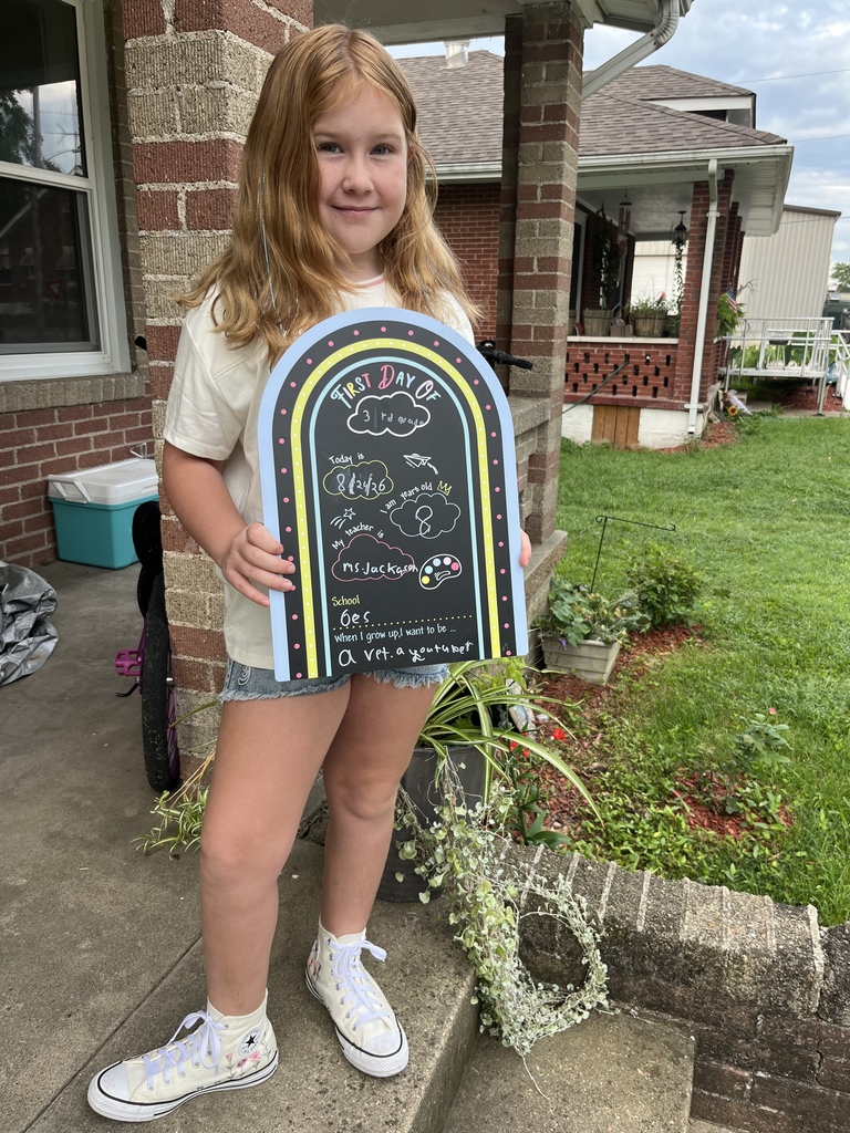 A girl with blond hair holds a rectangular sign with blue and white writing in front of a house.