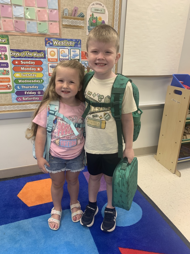 Two children, a boy and a girl, stand in a classroom. They are both wearing backpacks and smiling.