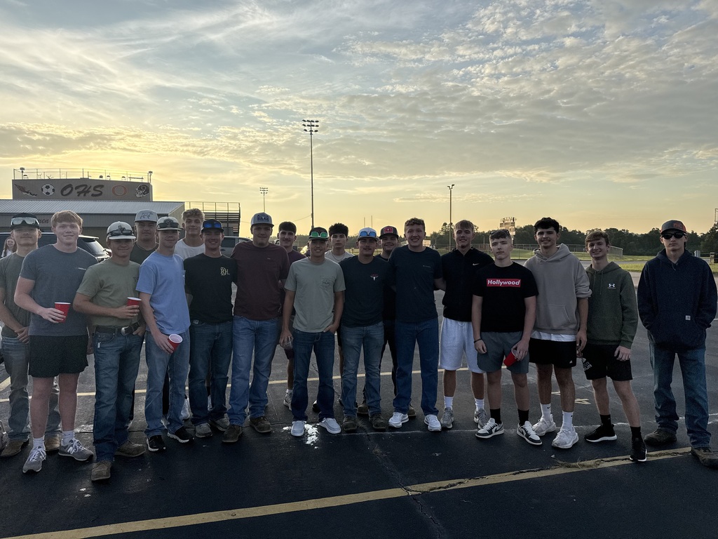 A group of people stands together in a parking lot, some wearing hats, at sunset.