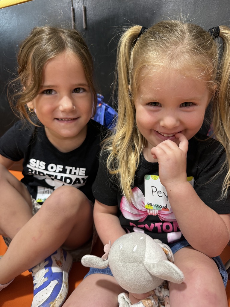 Two girls in black T-shirts sit close together. One holds a stuffed animal. They both smile and look at the camera.