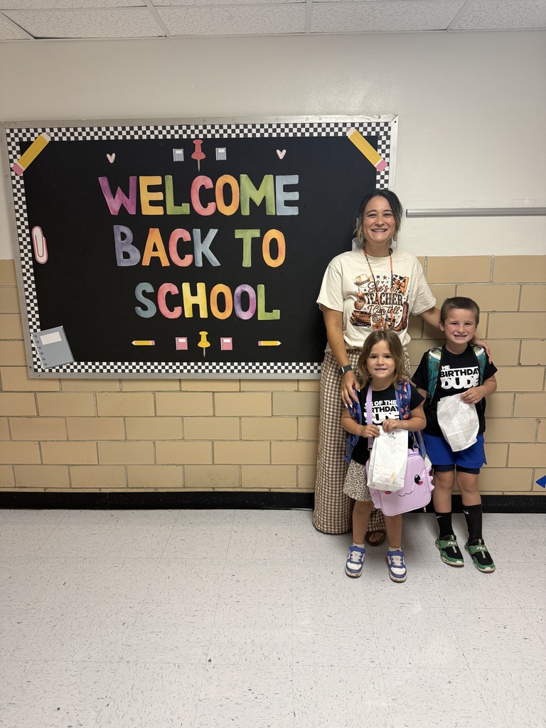 A woman and two children stand in front of a chalkboard with colorful writing, smiling.