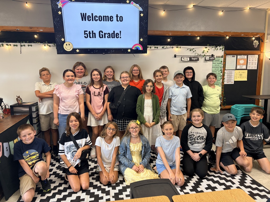 A group of young people, likely students, pose in a classroom. They sit or stand on a patterned floor.