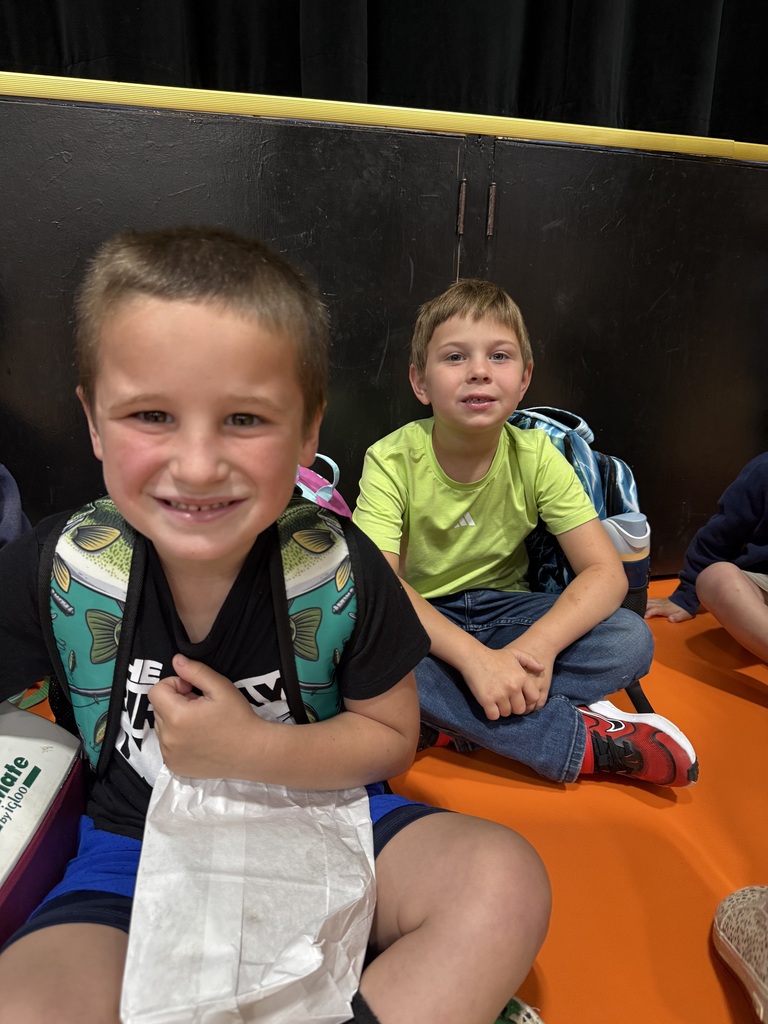 Two children sit on a padded floor, one holding a plastic bag, both wearing backpacks. A black wall and orange floor are behind them.