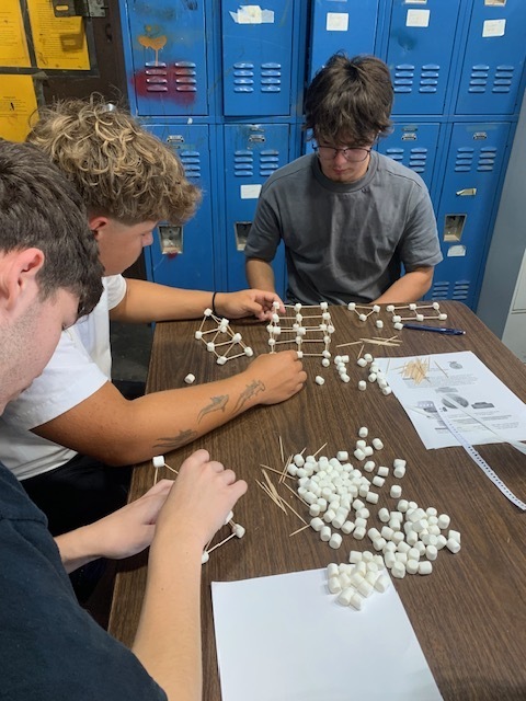 Three students work on a model with marshmallows and toothpicks at a table in a locker room.