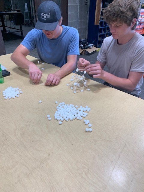 Two individuals work on a table, surrounded by small white beads, in a setting with storage boxes and a bench.