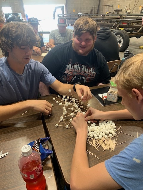 Three people in a workshop, two seated at a table constructing a structure with toothpicks and marshmallows, another watching. Background shows a vehicle.