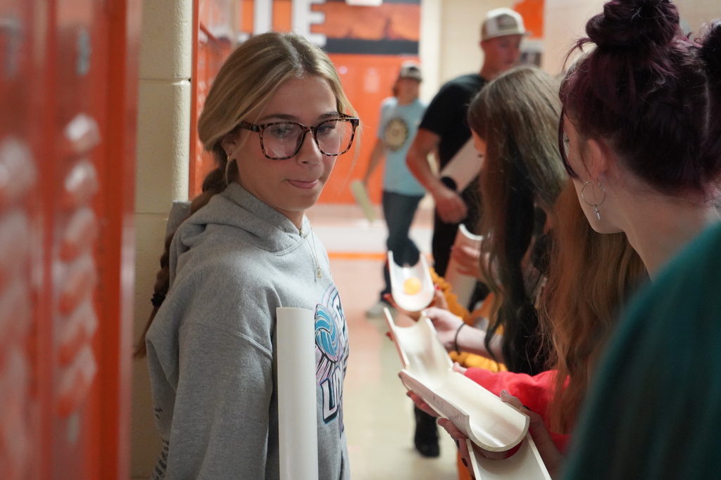 A woman in glasses stands in a hallway with a group of women, one holding an open box.