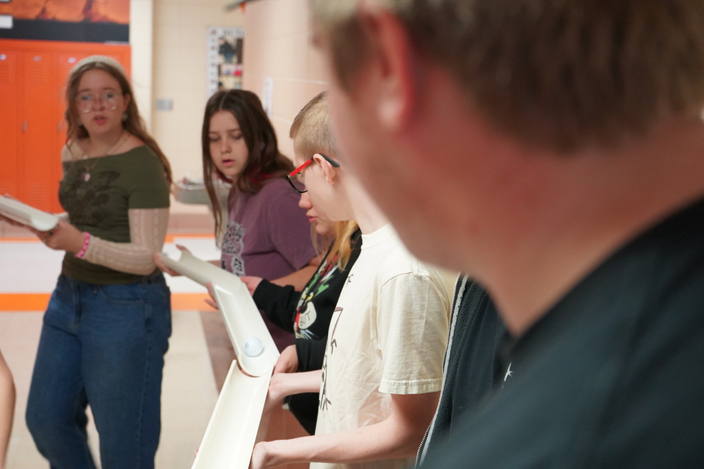 Five people, holding books and looking downward, are standing in a hallway. One person has glasses.