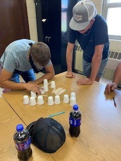Two people are working on a table with cups and bottles, one bending over and the other standing.