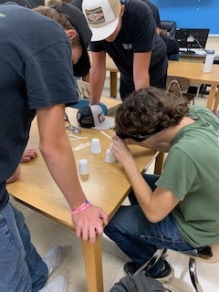 Students work on a table, wearing sunglasses and handling small objects, with two others standing nearby.
