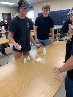 Three individuals in black t-shirts perform a cup stacking activity on a table, with additional people in the background.
