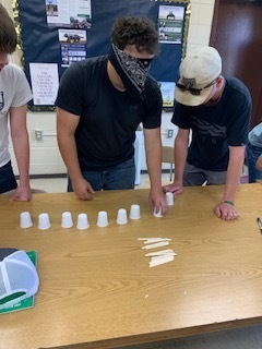 Three individuals in a classroom arrange small cups on a table. Two wear black shirts; one has a bandana, and the other has a hat.