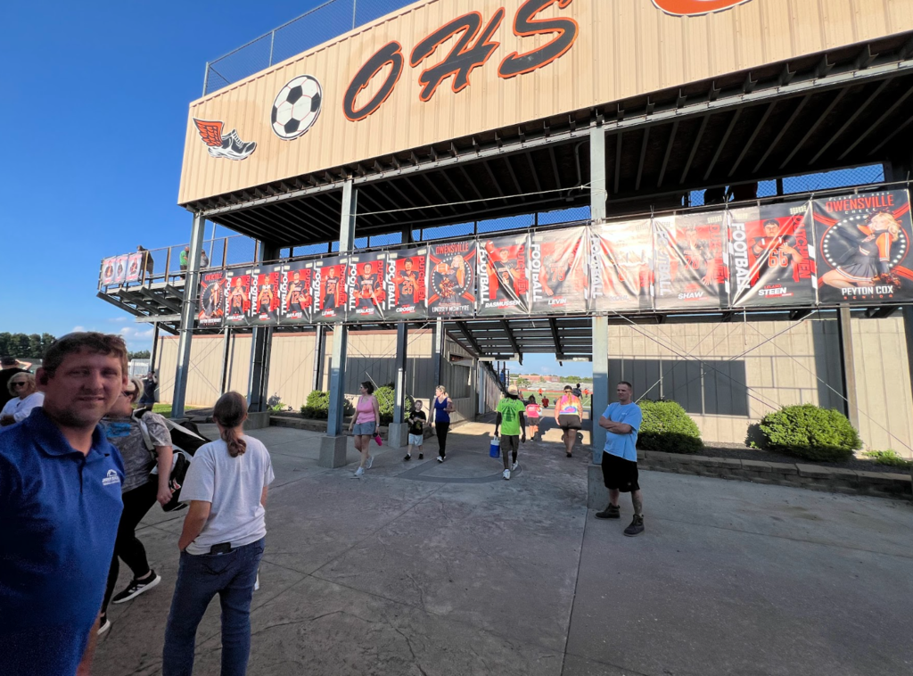A man in blue stands with others in front of a sports facility with "045" on it.