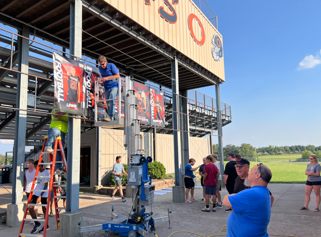 A group of people work on an outdoor structure with banners, using ladders and a lift.