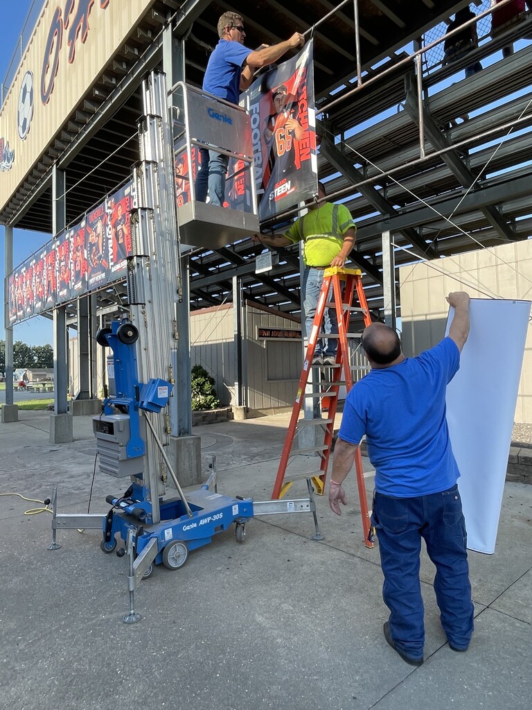 Workers are at a stadium, one using a ladder, another on a platform, and another holding a large white sheet.