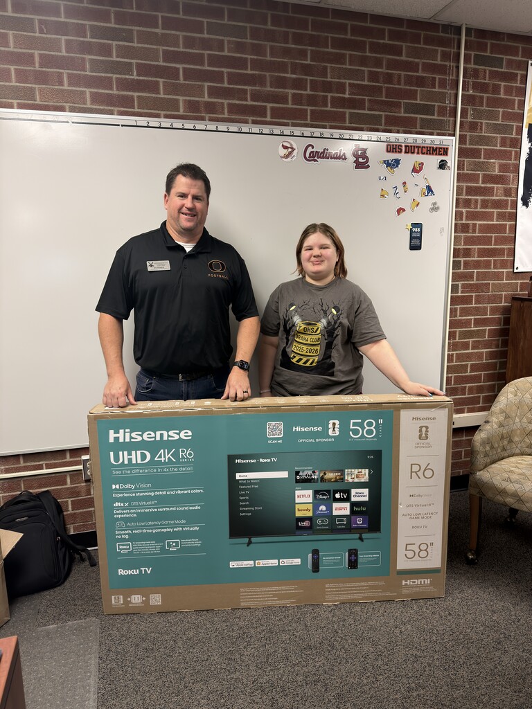 Two people stand behind a cardboard box with a large television label, standing in a room with brick walls.