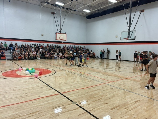 A basketball court with a crowd of people watching, a person throwing a ball, and others on the court.