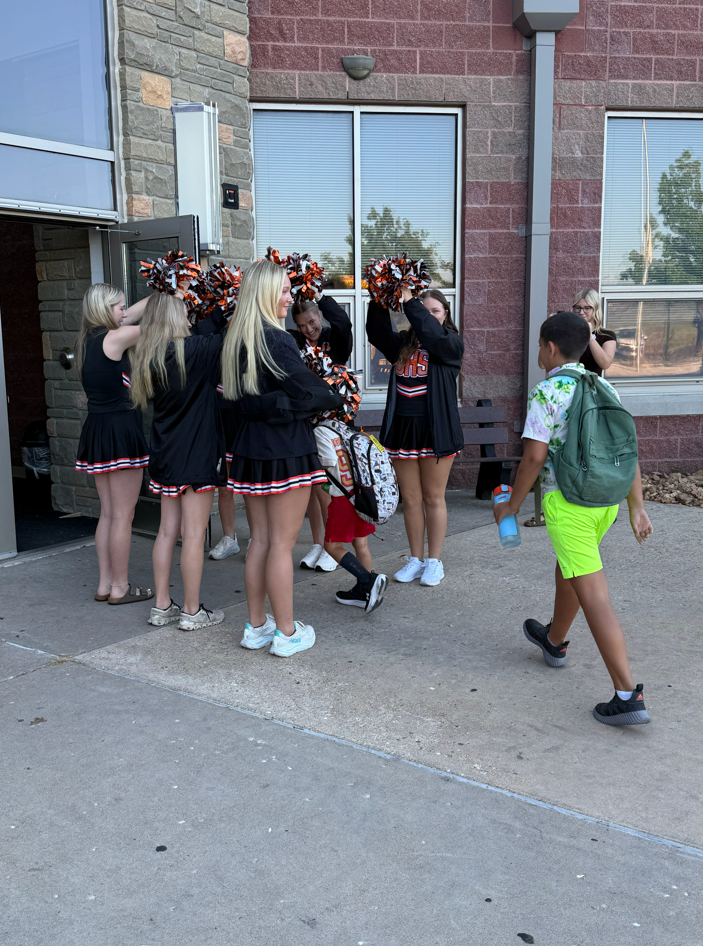 Cheerleaders stand near a building entrance, holding pom-poms. One student approaches, wearing a backpack.