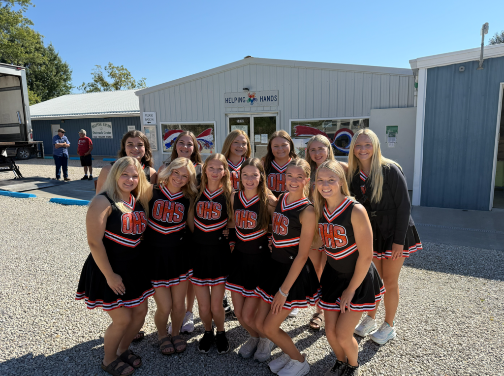 A group of cheerleaders in black uniforms with "HS" on them pose for a photo outside a building.