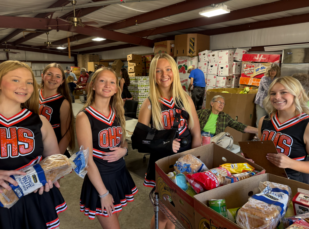 Five young women in cheerleading uniforms standing with boxes of food in a warehouse.